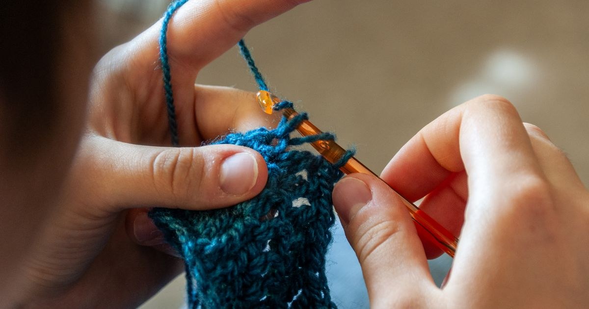 Hands crocheting a blue yarn piece with a crochet hook.