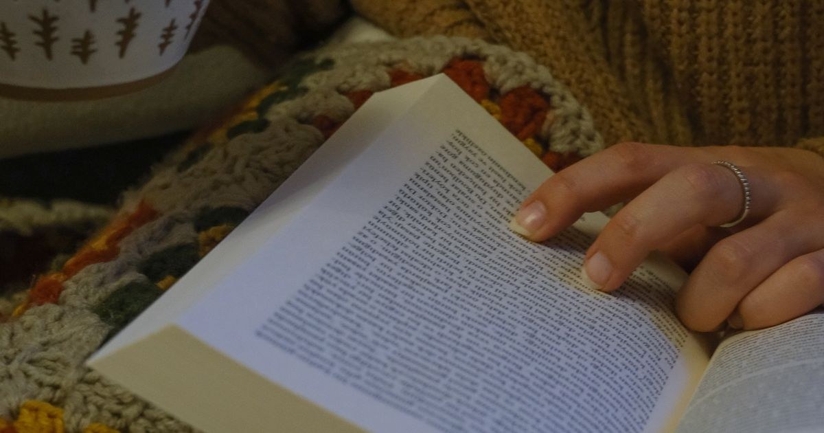 A cozy scene of a person holding a book and coffee mug, wrapped in a colorful blanket.