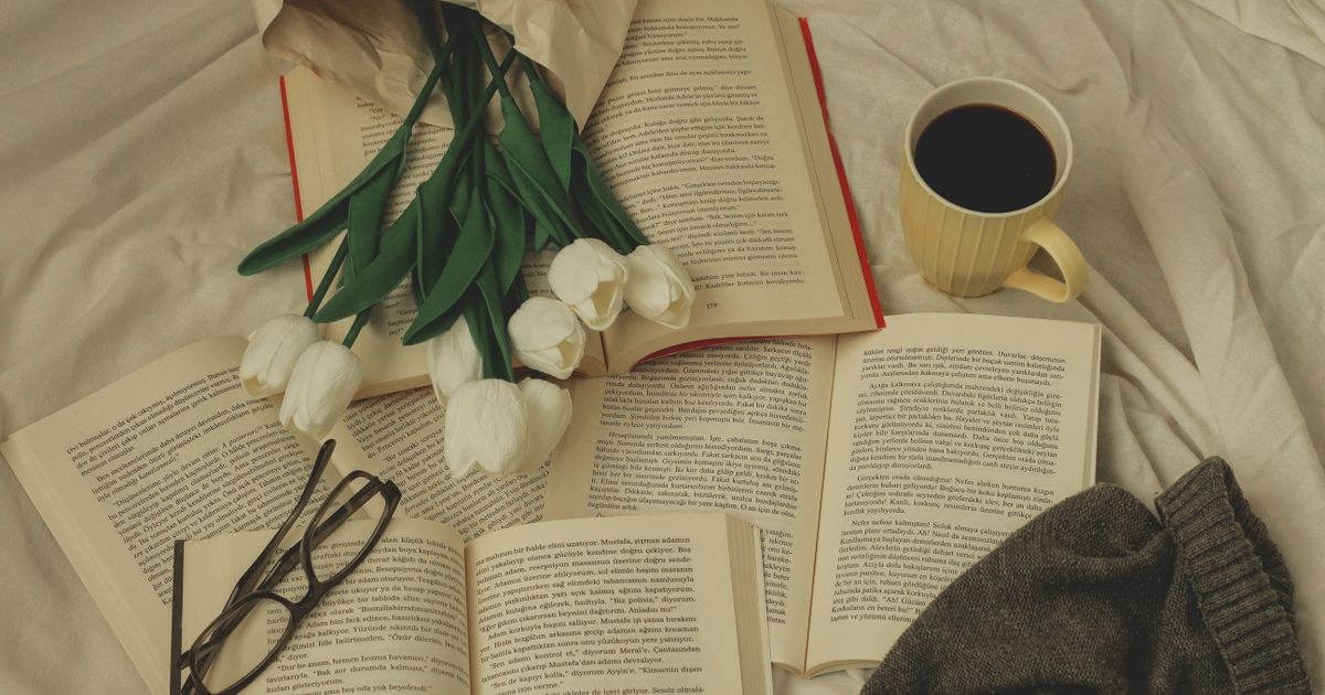 A relaxing setup with books, white tulips, a coffee mug, and reading glasses on a bed.