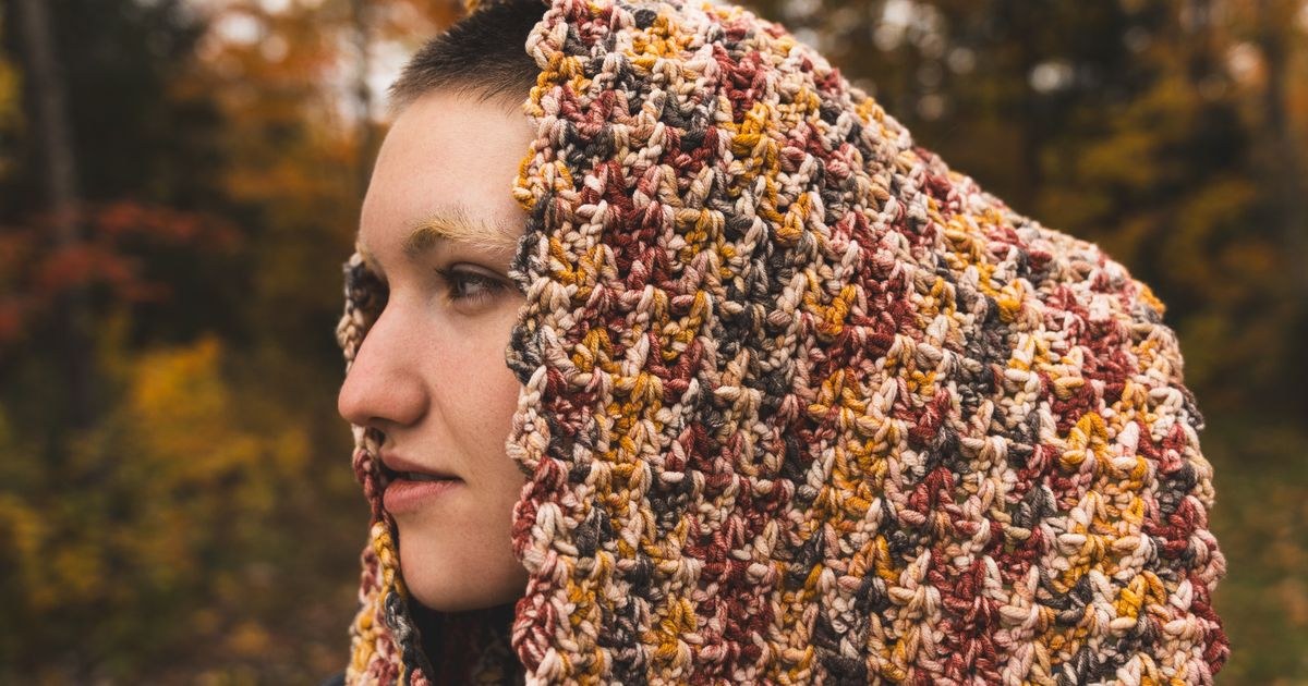 A young adult wearing a colorful scarf stands outdoors surrounded by autumn foliage.