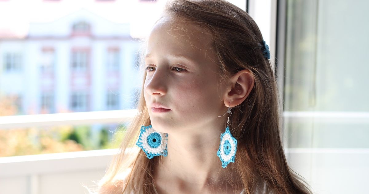 Portrait of a young girl with blue crochet earrings, gazing thoughtfully outdoors from a balcony.