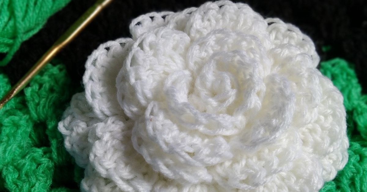 A close-up of a white crochet flower resting on a green crochet background with yarn and a needle.