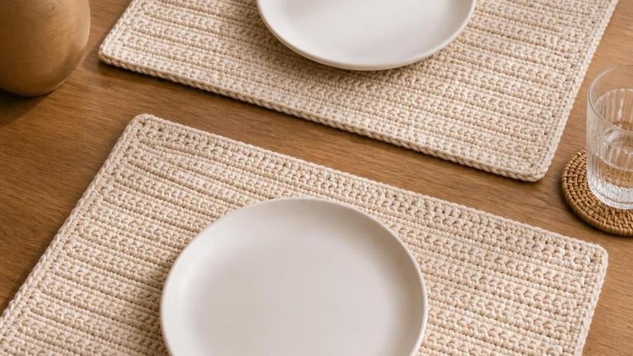 Two rectangular textured beige crochet placemats set on a wooden dining table with plates on top.