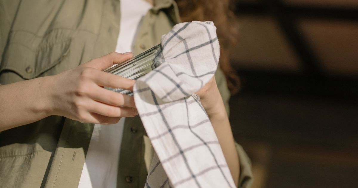 A person drying a clean glass with a white checkered dish towel indoors.