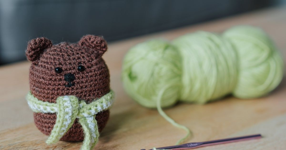 Closeup of small dark brown crochet toy bear and crochet next to light green threads on wooden table in bright room on blurred background