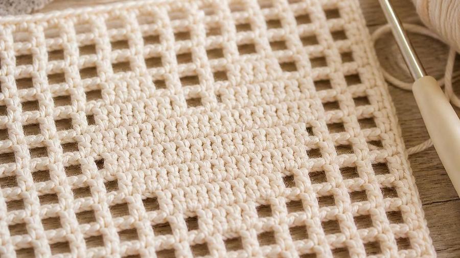 Close-up of a filet crochet swatch showing an open mesh grid pattern in cream cotton yarn, with a crochet hook at the edge.