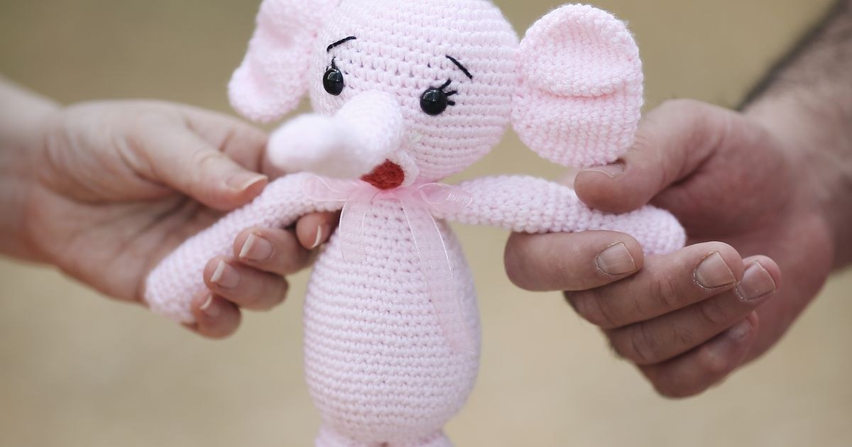 A pink crochet elephant amigurumi toy with safety eyes, held between two hands.