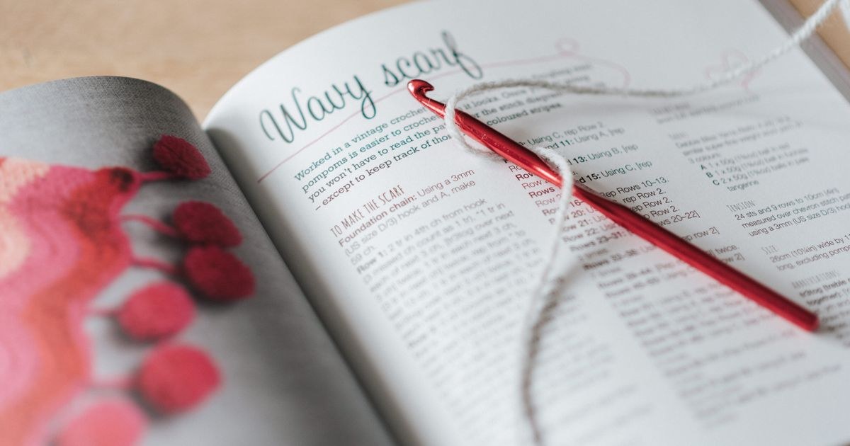 From above of red crochet needle and gray snaking thread on opened magazine on table in bright room