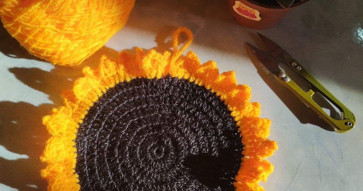 A hand-knitted sunflower surrounded by yarn, scissors, and a potted plant under daylight.