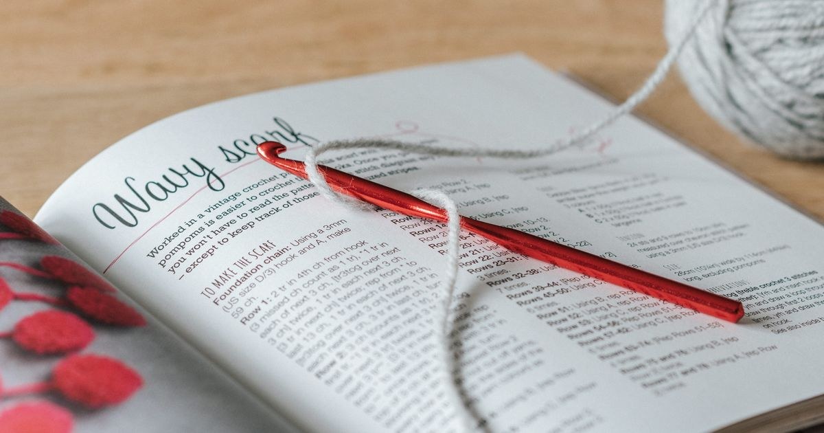 A ball of gray yarn with a crochet hook and a craft magazine on a wooden table, checking a pattern's gauge.