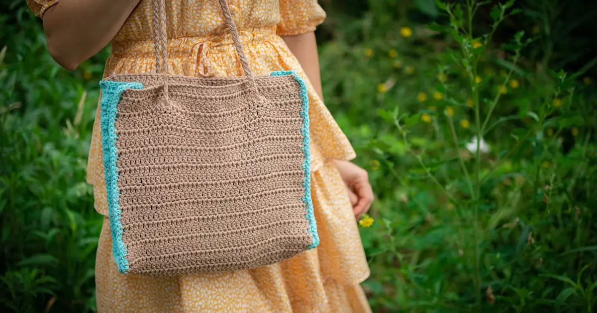 A woman in a yellow dress holding a crochet bag in a lush green outdoor scene.